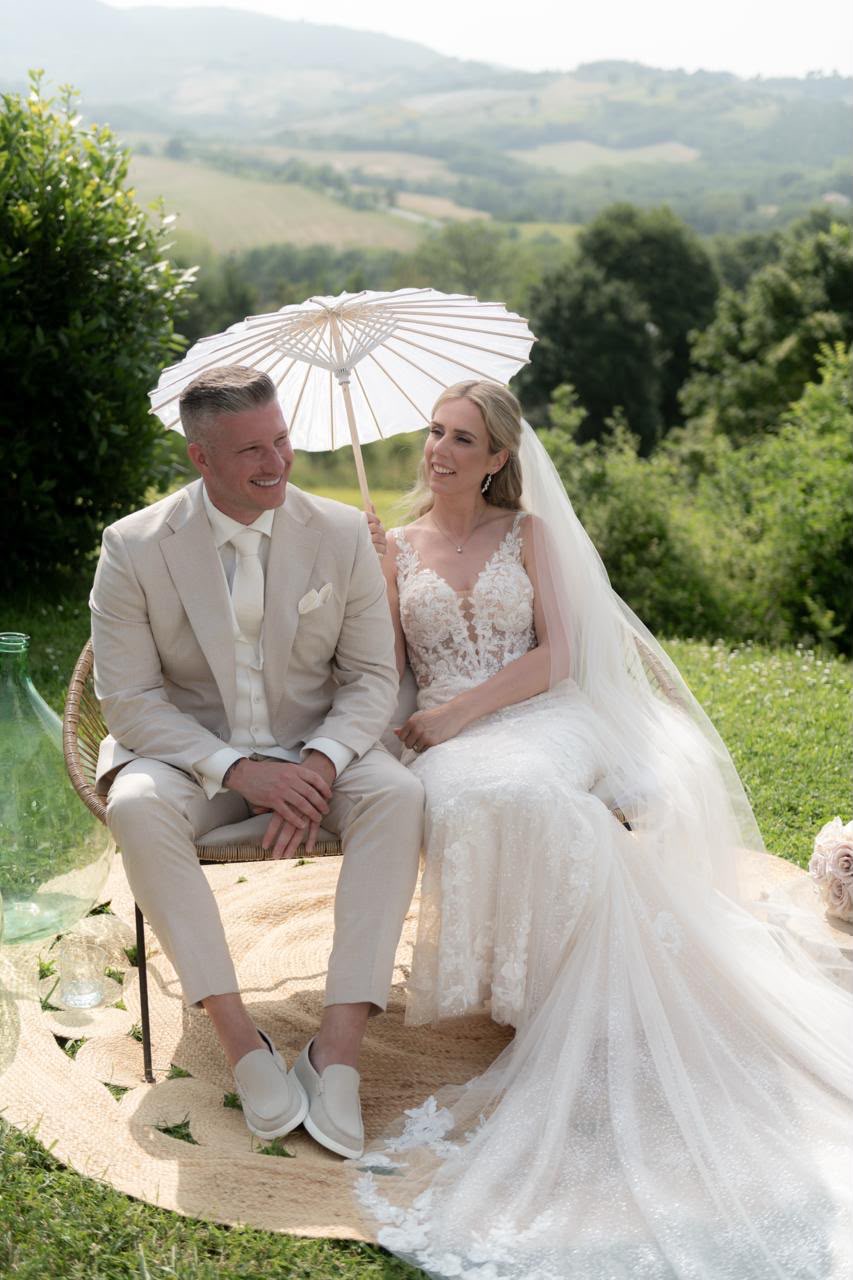Bruidspaar zit lachend naast elkaar in het groen, onder een witte parasol
